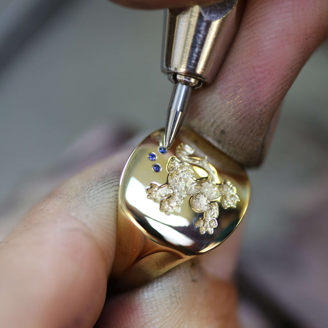 A jeweller is setting a blue sapphire on a yellow gold ring with a seal engraving of a dragon and two more blue sapphires on top.